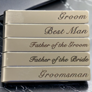 set of 5 tie clips on a black marble background reading 'Groom', 'Best Man', 'Father of the Groom', 'Father of the Bride' and 'Groomsman'.